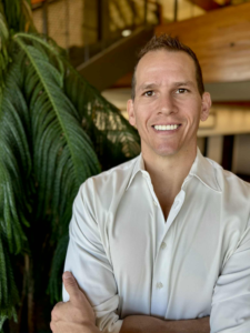 Dr Joe Heinecke, DC. Middle-aged white male in a white button-up shirt standing in front of green tree in office building.